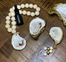 Load image into Gallery viewer, Gold glided oyster attached to a white wooden white blessing beads loop holding white Moroccan selenite crystals white a black Anuket diffusing oil bottle resting on them displayed on a wooden background