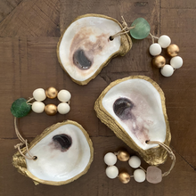 Charger l'image dans la galerie, Three gold glided oyster dishes attached to white and gold wooden blessing beads small loops two of them with a green stone and one with a light pink stone on a wooden background