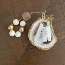 Charger l'image dans la galerie, Gold glided oyster dish holding white Moroccan selenite stick attached to a white and gold wooden blessing bead loop and a light pink stone loop displayed on a wooden background