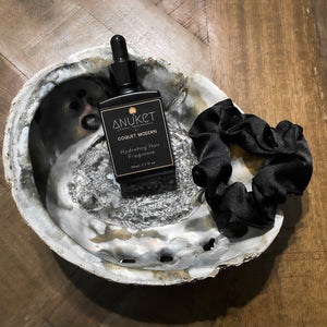 Hydrating hair fragrance bottle and black silk scrunchie displayed on a mother pearl dish on a wooden background