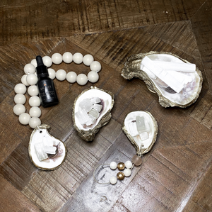 Four gold glided oyster holding Moroccan selenite crystals with one of them attached to white wooden white blessing beads and a diffusing oil bottle resting on them and one with a gold and white wooden bead and beige stone loop on a wooden background