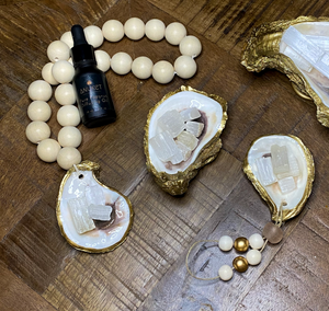 Pure Sakkara diffusing oil bottle laying on a white wooden blessing beads loop attached to gold glided oyster holding white selenite crystals with 3 other gold glided oysters displayed on a wooden background