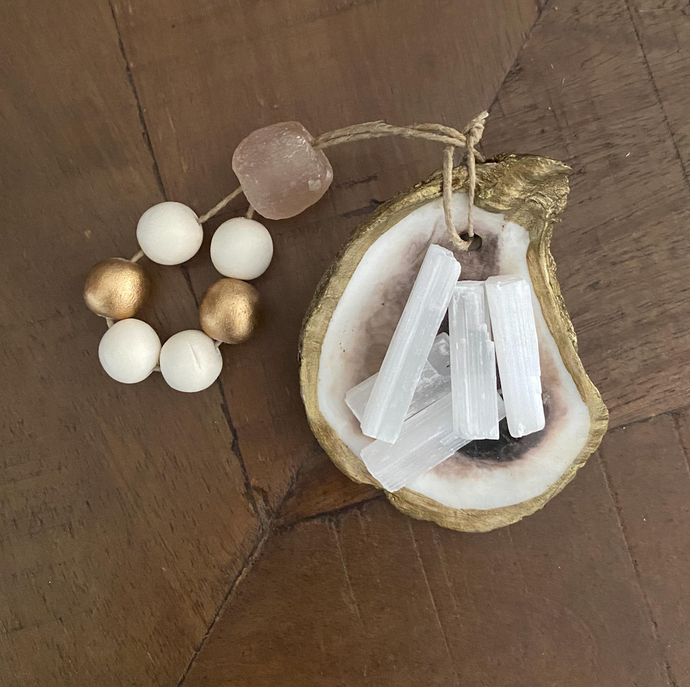 Gold glided oyster dish holding white Moroccan selenite stick attached to a white and gold wooden blessing bead loop and a light pink stone loop displayed on a wooden background