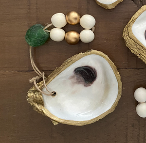 Gold glided oyster dish attached to a gold and white wooden blessing beads with a green stone small loop on a wooden background with small accents on two other gold glided oyster dishes